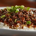 Mongolian ground beef served over steamed white rice, topped with green onions and sesame seeds, on a wooden table.