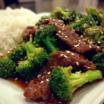 A plate of Chinese Beef and Broccoli stir-fry with beef strips and broccoli served with white rice, garnished with sesame seeds.