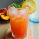 A chilled glass of peach lemonade with ice, mint, and a lemon slice, next to fresh peach slices on a wooden table.
